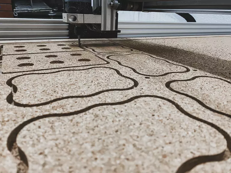 La imagen muestra una máquina de corte CNC trabajando en una hoja de material, creando varias formas y patrones. Esta escena es representativa del proceso de "prototipado", donde se utilizan tecnologías avanzadas para fabricar modelos y piezas de prueba.