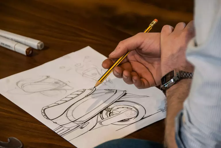 La imagen muestra una mano sosteniendo un lápiz mientras realiza un dibujo técnico de una motocicleta en una hoja de papel. Sobre la mesa se ven otros bocetos y algunos marcadores. Esta escena representa el "diseño industrial", donde un diseñador conceptualiza y desarrolla productos mediante dibujos y planos técnicos.