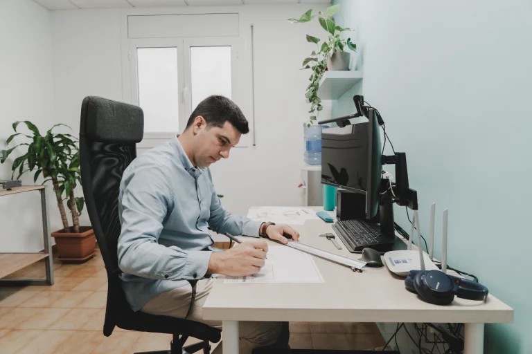 La imagen muestra a un hombre trabajando en un escritorio en una oficina. El hombre está enfocado en un dibujo técnico, utilizando una regla y un lápiz. En el escritorio hay una computadora, auriculares y varios instrumentos de trabajo. Al fondo se observan plantas y una estantería con más objetos. Esta escena ejemplifica el "diseño industrial y desarrollo de producto", donde el profesional se dedica a crear y perfeccionar diseños técnicos que serán utilizados en la producción de nuevos productos.