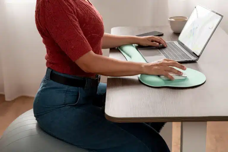 La imagen muestra a una persona trabajando en una computadora portátil en un escritorio, sentada sobre una pelota de ejercicio y utilizando un reposamuñecas ergonómico para el teclado y el mouse. Esta configuración destaca la importancia de la "ergonomía" en el entorno de trabajo