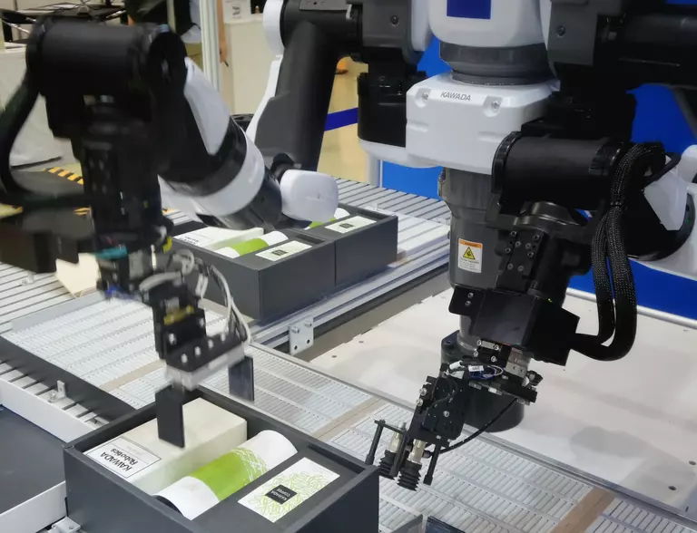La imagen muestra un sistema automatizado de robots trabajando en una línea de ensamblaje, manipulando y organizando productos en cajas. Este proceso ilustra el concepto de "fabricación flexible", donde la tecnología robótica permite la adaptación rápida a diferentes tareas de producción.